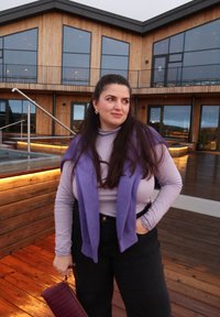 Lila gerippter Pullover über einem enganliegenden helllila Oberteil, kombiniert mit hoch taillierten schwarzen Hosen. Hält eine bordeauxrote Clutch. Hintergrund aus Holzdeck.