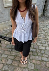 Λευκή μπλούζα peplum με κεντημένα λουλούδια και κορδόνια, συνδυασμένη με μαύρες βερμούδες και σαγιονάρες. Φοράει κολιέ με κόκκινες χάντρες. Χαλαρό εξωτερικό περιβάλλον.