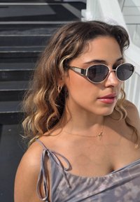 Slnečné okuliare s oválnymi čiernymi sklami, striebornými doplnkami a elegantným dizajnom. Nosené osobou s vlnitými vlasmi a jemným sivým topom s viazaním na remienky.