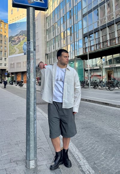 Camisa blanca ligera de manga larga sobre una camiseta gráfica gris; pantalones cortos grises; botas negras con cordones. Entorno urbano con edificios modernos.