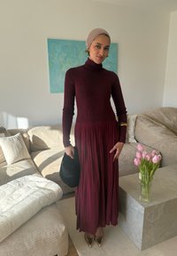 Femme portant une robe à col roulé bordeaux et un foulard beige, debout dans un salon, tenant un chapeau noir à côté d'un vase de tulipes roses.
