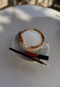 Witte koffie mok met schuimige cappuccino en gouden rand op een schotel, vergezeld van twee lipproducten: een potlood en vloeibare lipkleur.