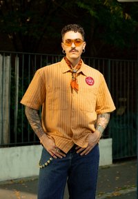 Uomo con capelli ricci, baffi e tatuaggi indossa una camicia a righe arancioni, una sciarpa arancione, occhiali da sole e jeans blu, in piedi all'aperto con le mani in tasca.