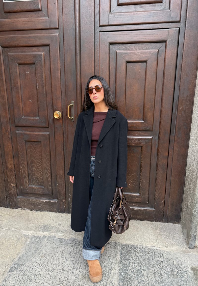 Schwarzer Wollmantel, brauner Pullover, blaue Jeans mit umgeschlagenen Bündchen, braune Plateaustiefel, hält eine braune Handtasche, steht vor einer Holztür.