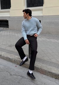 Maglione a coste azzurro chiaro, pantaloni neri, mocassini neri e calzini bianchi. Seduto sul bordo del marciapiede con pavimentazione testurizzata.