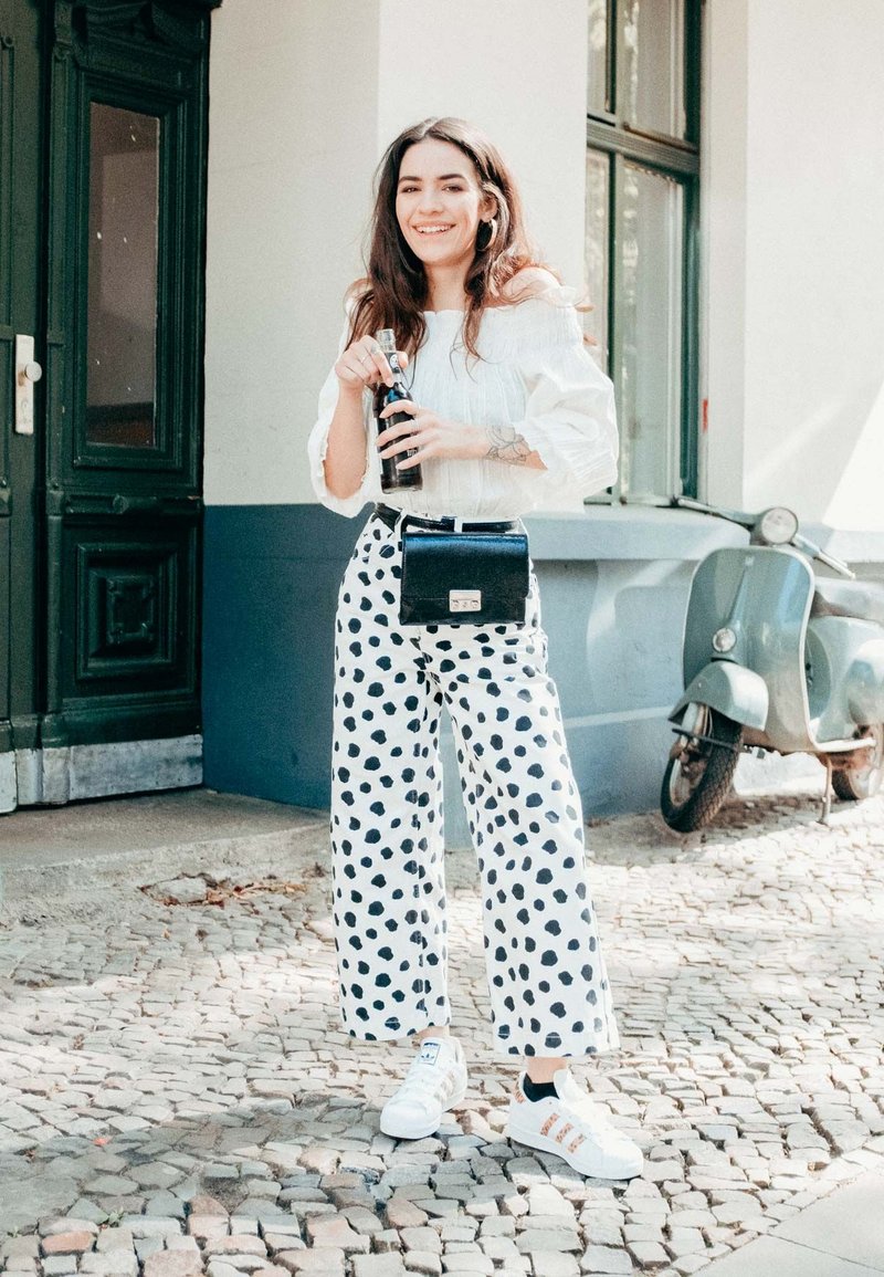 Fehér, vállon lecsúszó blúz, fekete pöttyös, széles szárú nadrág, fekete keresztpántos táska és fehér sportcipő. Macskaköves talaj és zöld ajtó háttér.