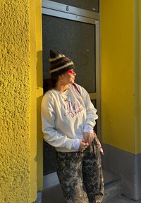 Sweat-shirt blanc avec lettres rouges, pantalon camouflage, bonnet tricoté rayé, et lunettes de soleil rouges. Fond de mur gris et jaune. Lumière naturelle.