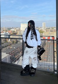 White hoodie with black accents and bows, distressed white trousers, and large black boots. Black handbag with chain detail. City skyline backdrop.