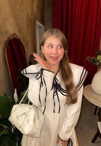 Hvid kjole med sorte accenter, lange ærmer bundet ved skuldrene. Matchende hvid håndtaske med tekstureret design. Baggrunden har røde gardiner.