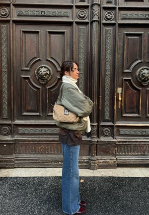 Bolso de hombro de lona beige con patrón clásico marrón, herrajes plateados y forma curva, sostenido contra un fondo de madera oscura texturizada.