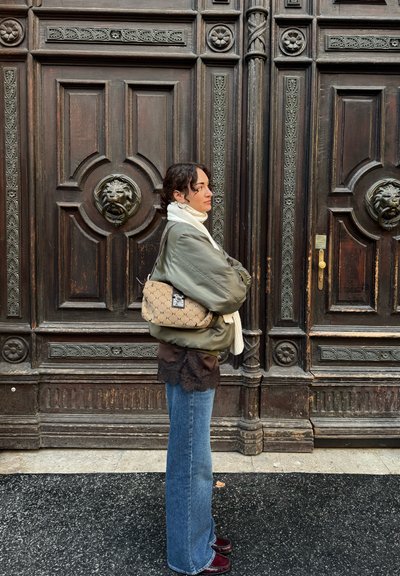 Bolso de hombro de lona beige con patrón clásico marrón, herrajes plateados y forma curva, sostenido contra un fondo de madera oscura texturizada.