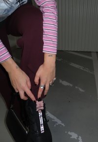 Personne se penchant, portant des manches rayées roses, un pantalon bordeaux, des baskets noires avec une étiquette rose, et des bagues argentées aux doigts.