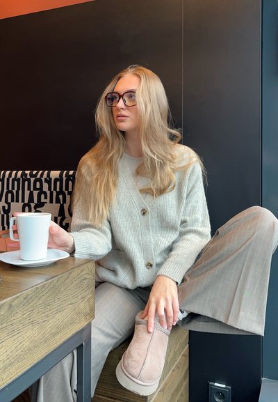 Cardigan de punto gris claro con botones, combinado con pantalones de cuadros beige y unas botas de ante rosa hasta el tobillo. Taza blanca sobre una mesa de madera.