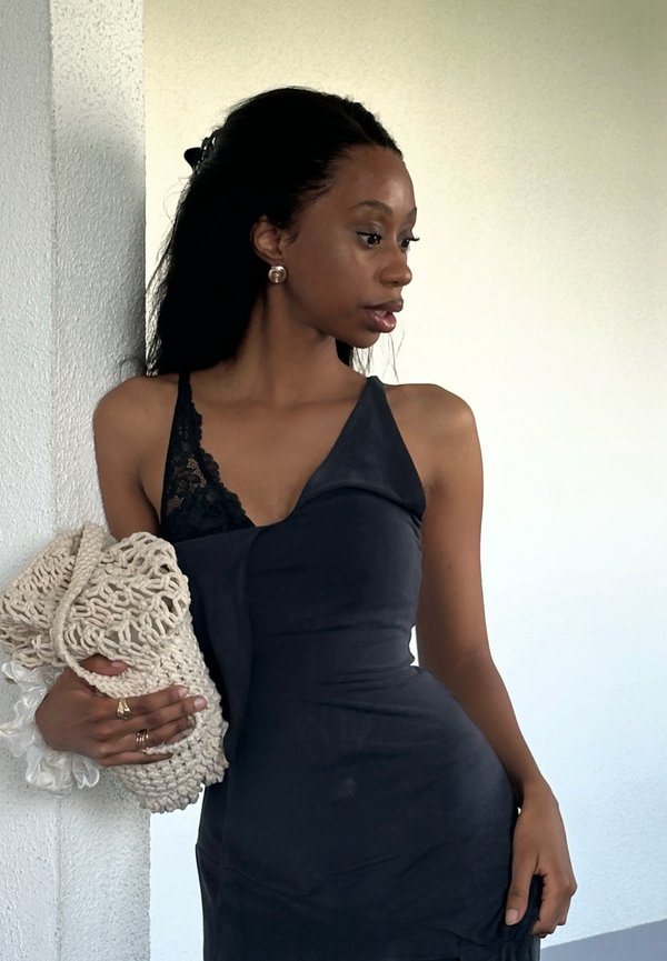 Femme en robe noire asymétrique tenant un sac tissé beige, debout contre un mur blanc texturé, regardant vers sa droite.
