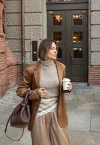 Blazer din suede maro peste un pulover bej cu guler înalt, eșarfă crem cu model și detalii argintii, pantaloni largi bej, ținând o cană de cafea, geantă de umăr maro.