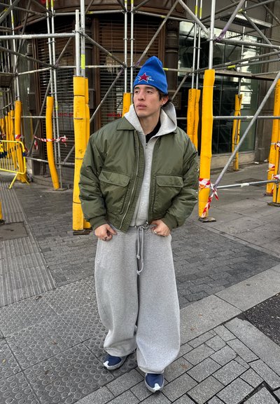 Joven con chaqueta verde oliva, sudadera con capucha gris, pantalones anchos grises, gorro azul y zapatillas, de pie en una acera urbana cerca de un andamio.