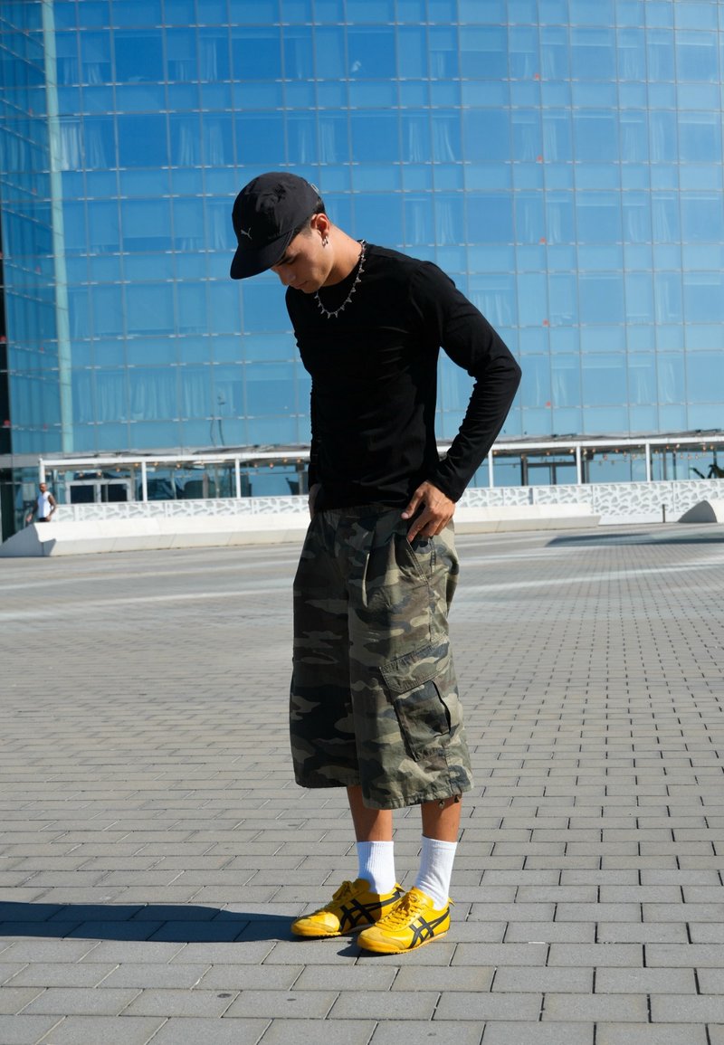 Jeune homme en manches longues noires, short camouflage, baskets jaunes et casquette noire debout sur une surface pavée avec un bâtiment en verre bleu derrière.
