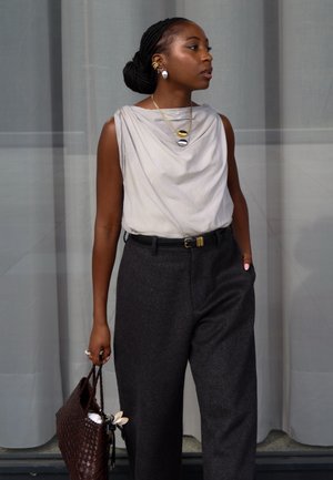 Mujer con cabello trenzado, vestida con una blusa ligera sin mangas y pantalones oscuros de tiro alto, sosteniendo un bolso marrón tejido, de pie frente a una cortina gris.