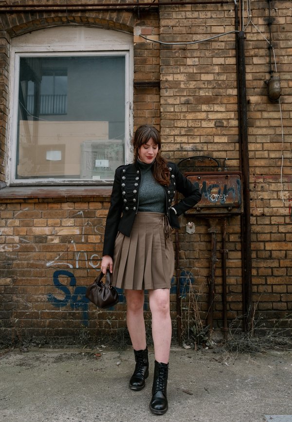 Mujer con cabello castaño que lleva una chaqueta negra, top gris, falda marrón plisada, botas negras y sostiene un bolso marrón, de pie junto a una pared de ladrillos.