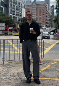 Camisola preta de manga comprida, calças cinzentas plissadas, segurando uma chávena de café, com óculos de sol e um relógio. Ambiente urbano ao fundo.