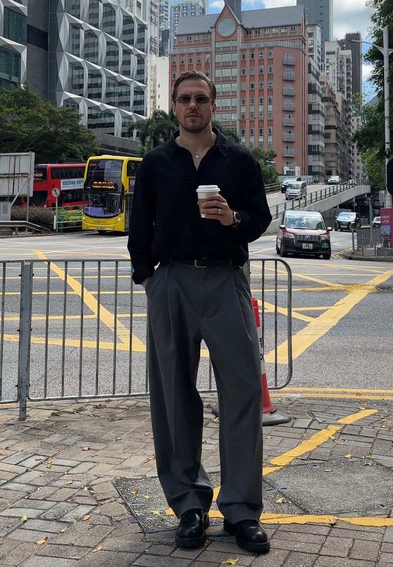 Camicia nera a maniche lunghe, pantaloni grigi plissettati, tiene una tazza di caffè, con occhiali da sole e orologio. Sfondo urbano.