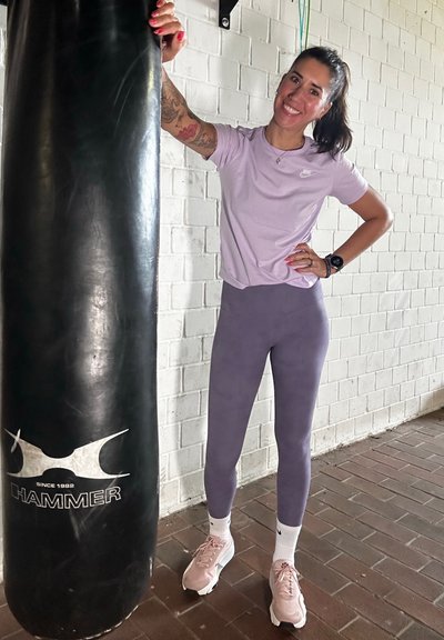 Camiseta atlética lila, leggings púrpura oscuro, zapatillas rosas. De pie junto a un saco de boxeo negro con el logo "HAMMER". Suelo texturizado.