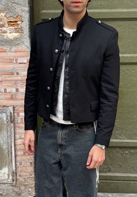 Veste courte noire avec boutons argentés, col montant et poches à rabat, portée sur une chemise blanche. Associée à un pantalon en jean foncé.