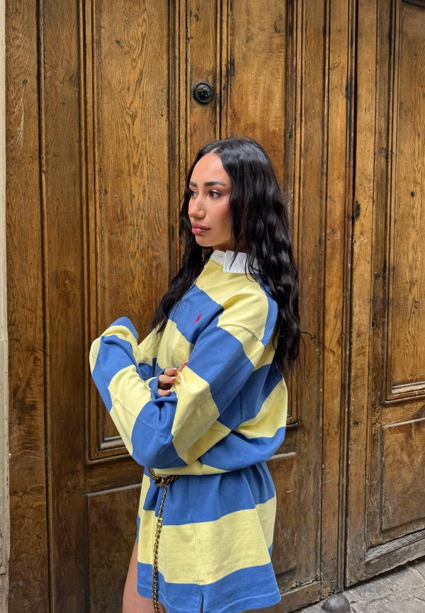 Femme aux longs cheveux noirs portant une chemise et une jupe rayées bleu et jaune, debout de profil devant des portes en bois à panneaux.