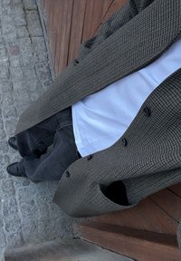 Manteau marron à motif pied-de-poule avec des boutons noirs, porté sur une chemise blanche et un pantalon noir. Chaussures noires, debout sur un pavé en pierre.