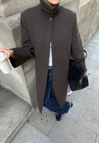 Bruine wollen jas van een wolmix met een hoge kraag en knoopsluiting. Gecombineerd met blauwe spijkerbroeken en een zwarte handtas. Beige koffiekop in de hand.