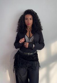 Cămașă neagră cu butoni, purtată peste un bralette cu model, fustă neagră cu ciucuri, bijuterii din argint și coliere din mărgele, pe un fundal luminos.