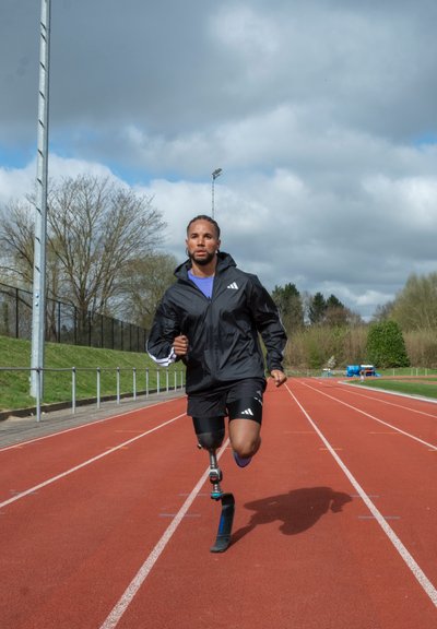 Corredor con chaqueta negra y camiseta morada en una pista roja, utilizando una pierna prostética de fibra de carbono, con cielos nublados de fondo.