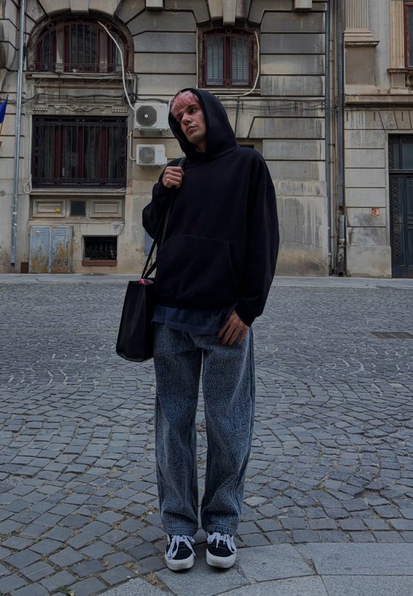 Jeune personne aux cheveux roses portant un sweat à capuche noir et un jean ample se tient dans une rue pavée devant un vieux bâtiment en pierre.