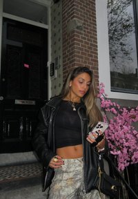 Czarna skórzana kurtka na czarnym krótki topie, spodnie moro, trzymająca telefon. W tle różowe kwiaty i strukturalna ceglasta ściana.