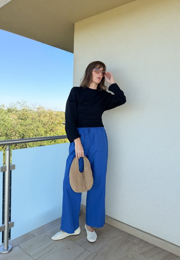 Femme portant des lunettes de soleil, un haut à manches longues noir, un pantalon bleu à jambes larges et des chaussures blanches, debout sur un balcon tenant un sac à main ovale beige.