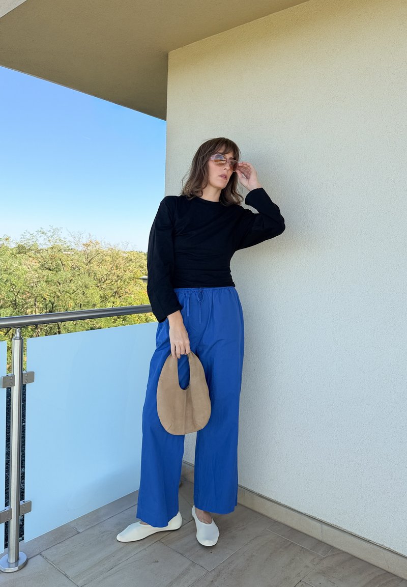Femme portant des lunettes de soleil, un haut à manches longues noir, un pantalon bleu à jambes larges et des chaussures blanches, debout sur un balcon tenant un sac à main ovale beige.