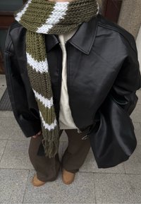 Giacca bomber in pelle nera, sciarpa lavorata a maglia spessa a strisce verde oliva e bianco, maglione crema, pantaloni marroni e scarpe slip-on color tan.