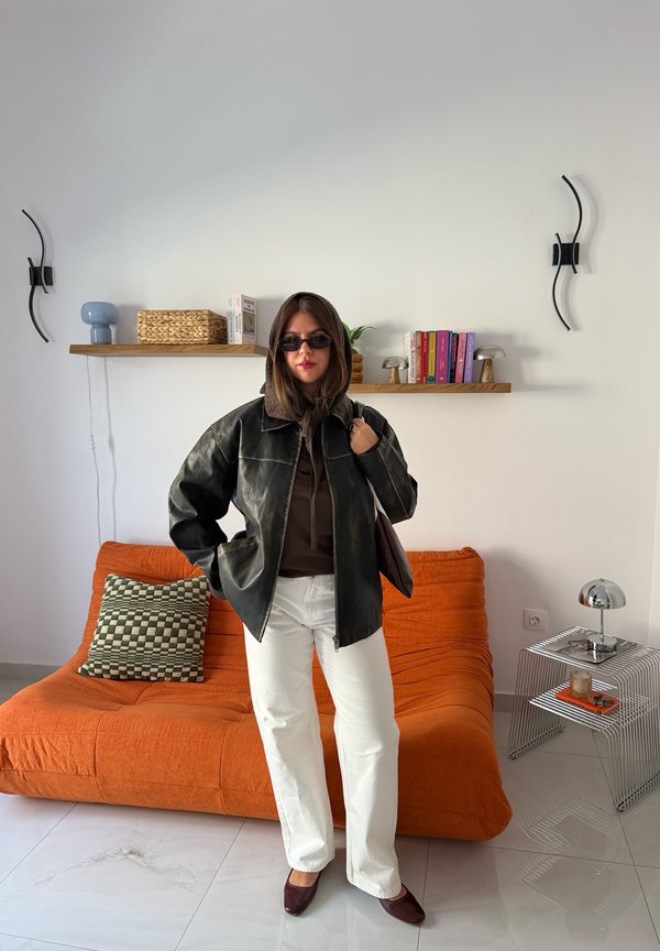Mujer con gafas de sol, chaqueta de cuero negra, sudadera marrón, pantalones blancos y zapatos marrones está delante de un sofá naranja en una habitación moderna.