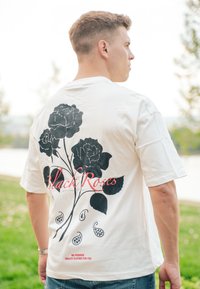 Vit kortärmad t-shirt med en stor svart ros grafik och röd text "Black Roses" på ryggen, tillsammans med dekorativa paisley-element.