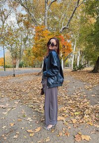 Melnā ādas bomber jakas, pelēkas platas bikses un melnbaltas svītrainas papēžkurpes; āra vide ar rudenīkiem lapām un kokiem.