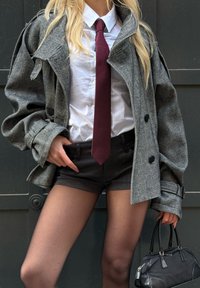 Cappotto grigio a spina di pesce con ampi risvolti, camicia bianca, cravatta a strisce bordeaux, pantaloncini neri e borsa nera. Texture liscia e vestibilità sartoriale.