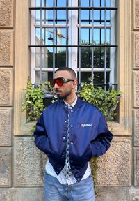 Giacca bomber in satin navy con bottoni a pressione, logo "ORIGINALS", abbinata a una camicia a quadri, jeans in denim e occhiali da sole rossi oversize.