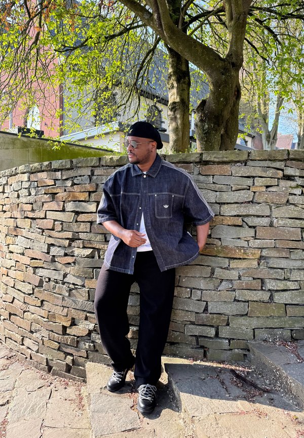 Homme portant des lunettes de soleil, une casquette noire, une chemise en denim foncé et un pantalon noir, appuyé contre un mur en pierre sous un arbre aux feuilles vertes.