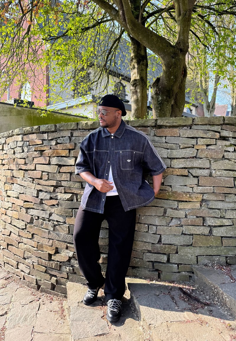 Mann mit Sonnenbrille, schwarzer Kappe, dunkelblauem Jeanshemd und schwarzer Hose lehnt an einer Steinmauer unter einem Baum mit grünen Blättern.