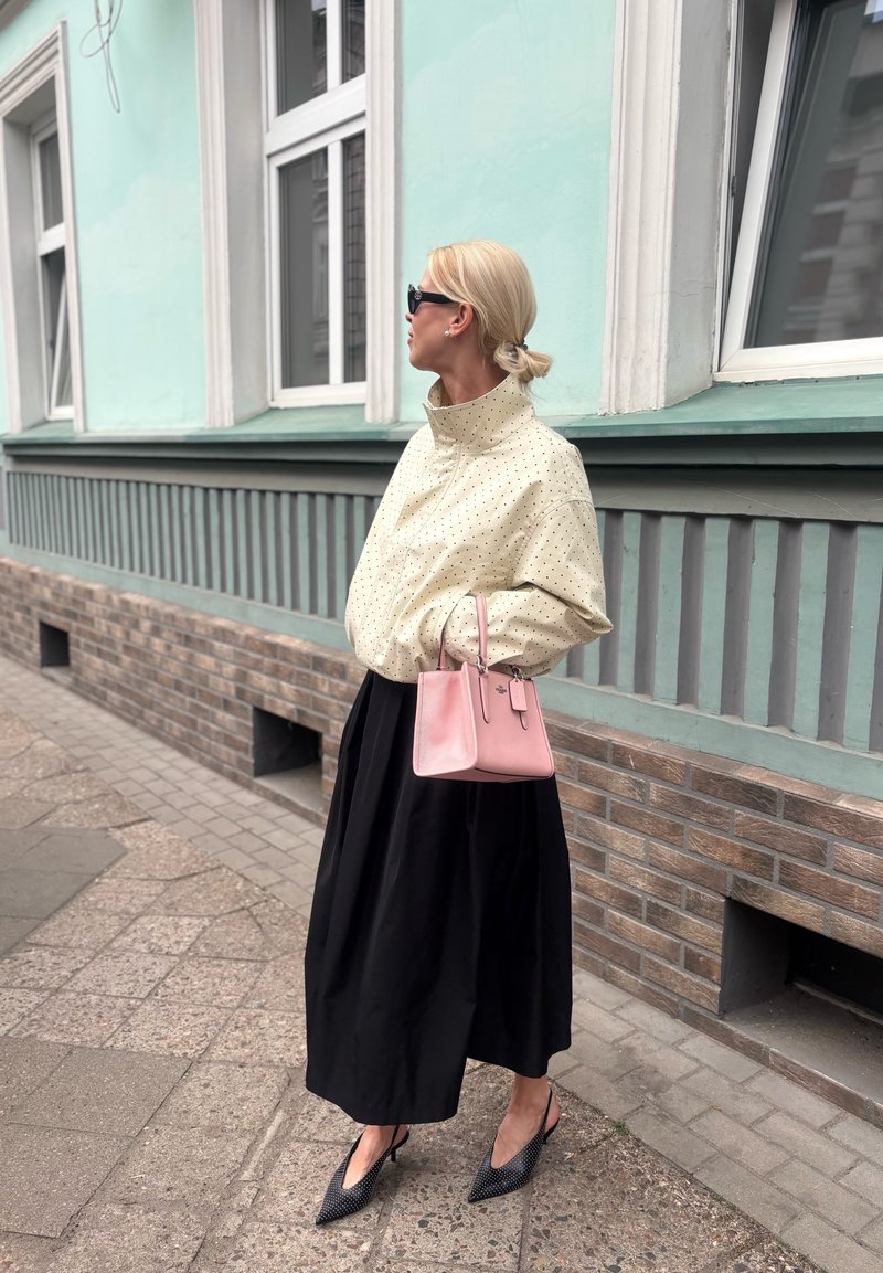 Femme aux cheveux blonds en chignon, portant des lunettes de soleil, une veste crème à pois, une jupe noire, des talons noirs, tenant un sac à main rose près d'un bâtiment bleu sarcelle.