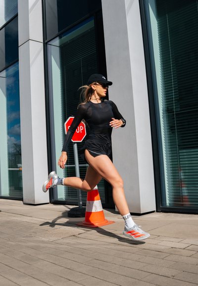 Mujer con ropa deportiva negra y gorra saltando sobre un cono de tráfico naranja en la acera de una ciudad cerca de un edificio de vidrio y una señal de stop.