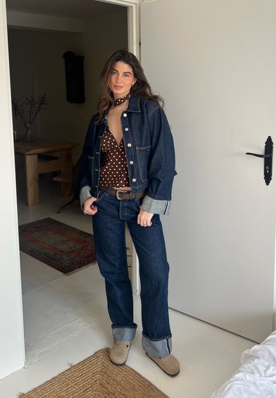 Chaqueta de denim con mangas enrolladas, blusa de lunares, jeans de cintura alta y zuecos beige. Colores neutros y corte clásico con un estilo informal.