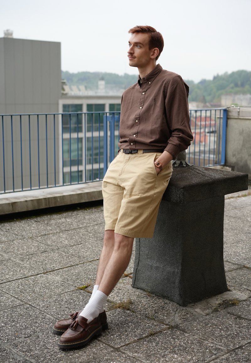Jeune homme en chemise marron et short beige appuyé sur une structure de toit, regard pensif avec des bâtiments de la ville et de la verdure en arrière-plan.