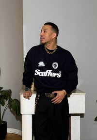 Fekete pulóver fehér grafikával és rózsamintával, laza fekete nadrággal, vastag övvel lánccal és arany kiegészítőkkel párosítva.