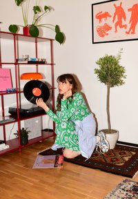 Vestido verde com padrão de flores brancas, segurando um disco de vinil preto. O ambiente apresenta uma estante vermelha aberta, plantas e um tapete colorido.
